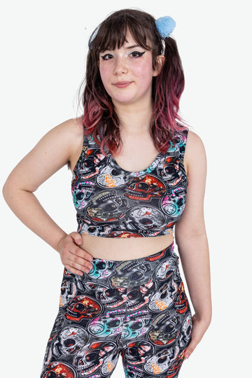 Emo Skulls active loungewear crop comfort top is black grey with red blue orange emo alternative music bands print. Worn with matching legging by an alternative model with brown and pink hair in bunches, with heavy winged eyeliner, posing with hands on hips. Made sustainably in the UK by Wilde Mode.