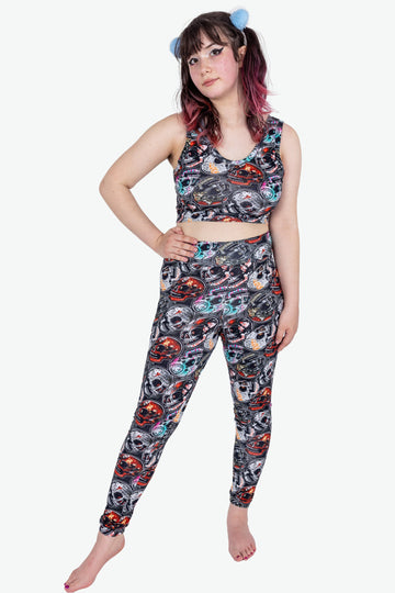 Emo Skulls active loungewear leggings are black grey with red blue orange emo alternative music bands print. Worn by an alternative model with brown and pink hair in bunches, with heavy winged eyeliner, posing with hand on hip. Made sustainably in the UK by Wilde Mode.
