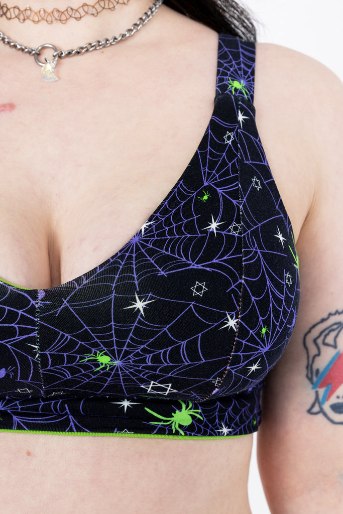 Close up of Tattooed model with green hair wearing Purple Spider Comfort Lounge Bra. The print has a black background with purple spiderwebs, green spiders and white starts all over. Close up shows the pattern.