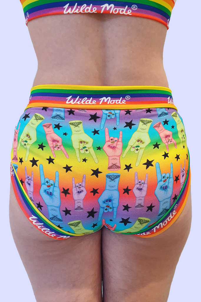 Model wearing Hail to the King Comfort Briefs. The print has a rainbow gradient background with blue, red, green and purple hands with red nails and moth tattoo on wrist all over doing rock and roll sign and black stars all over. The briefs have Wilde Mode rainbow banding. Model is posing with back to camera to show back of briefs.