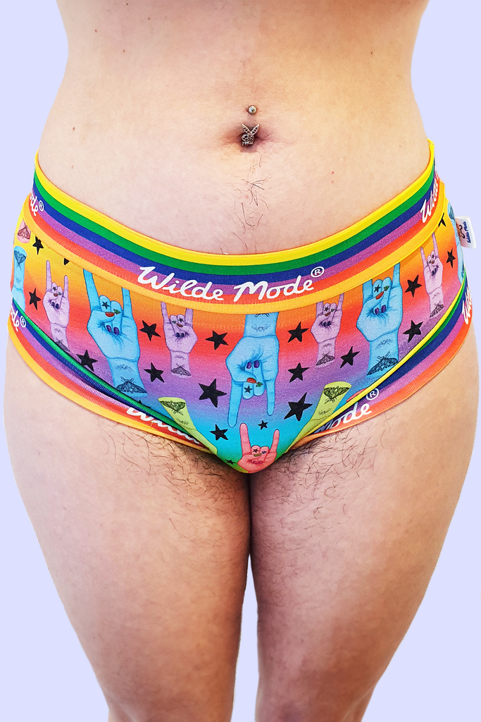 Model wearing Hail to the King Comfort Briefs. The print has a rainbow gradient background with blue, red, green and purple hands with red nails and moth tattoo on wrist all over doing rock and roll sign and black stars all over. The briefs have Wilde Mode rainbow banding. 