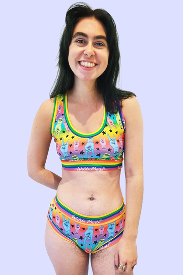 Model wearing Hail to the King Comfort Briefs. The print has a rainbow gradient background with blue, red, green and purple hands with red nails and moth tattoo on wrist all over doing rock and roll sign and black stars all over. The briefs have Wilde Mode rainbow banding. Model is smiling at camera with one hand by side and other behind back.