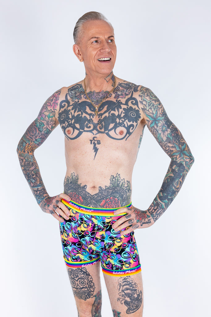Tattooed male model wearing Wilde Mode Neurodivergent Boxers shorts. The print features the rainbow infinity symbol on a multicoloured background with butterfly logo, the elastic hem is rainbow coloured with Wilde Mode in white writing. 