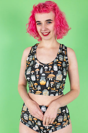 Lottie a tall white pink haired model is wearing a mushroom potions print on a black background two piece underwear set in a crop top and high waisted brief combo. They are looking at camera and smiling. The background is green.