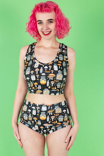 Lottie a tall white pink haired model is wearing a mushroom potions print on a black background two piece underwear set smiling at camera. The background is green.