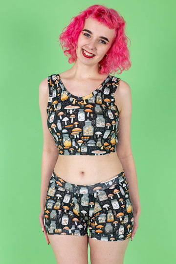 Lottie a tall white pink haired model is wearing a mushroom potions print on a black background two piece underwear set in a crop top and boxer short combo smiling at camera. The background is green.