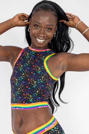 Model with long black ponytail wearing Rainbow Splat Racer Front Top. The print is a black background with rainbow coloured paint platters all over. The top has Wilde Mode rainbow banding. Model is smiling at the camera with hands in hair.