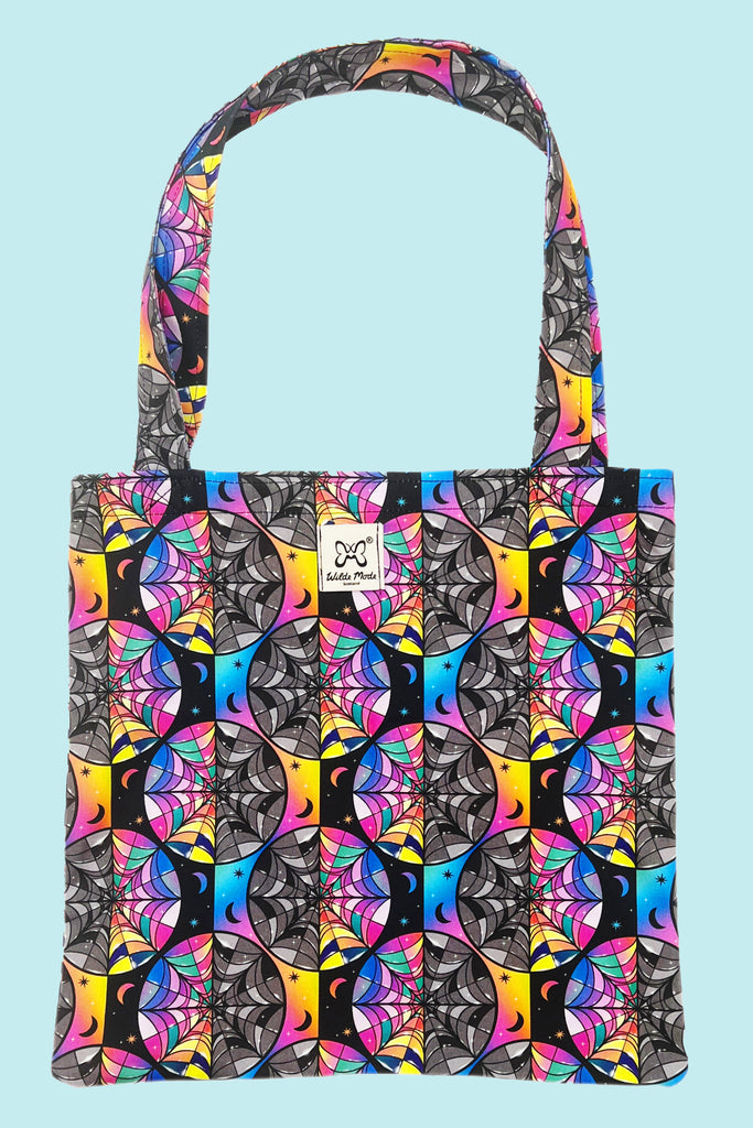 A black and rainbow tote bag with spiderweb shaped stained glass windows, half rainbow coloured and half black and white. There are various stars and moons dotted around the print.