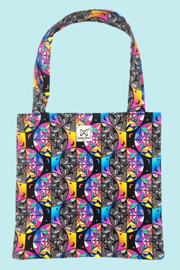A black and rainbow tote bag with spiderweb shaped stained glass windows, half rainbow coloured and half black and white. There are various stars and moons dotted around the print.