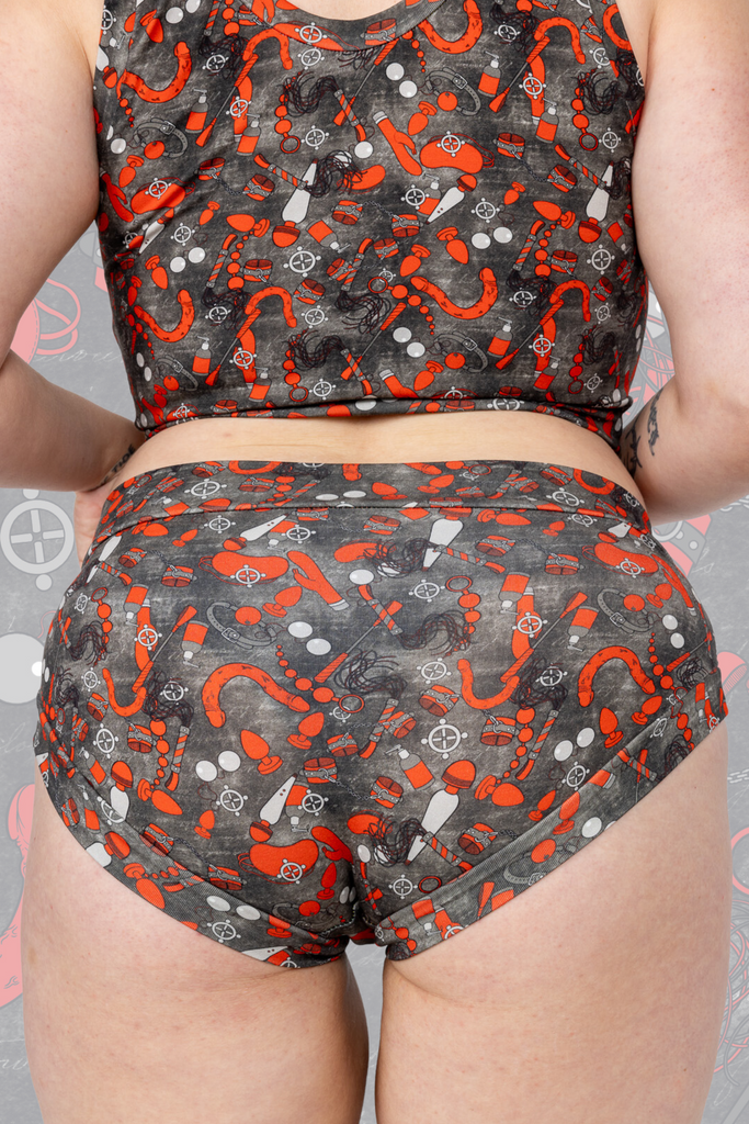 Model is wearing What’s Your Kink? 18+ Ultra Naughty Comfort Briefs paired with comfort top. The briefs are grey with various red sex toys all over. Model is posing with bum to camera.