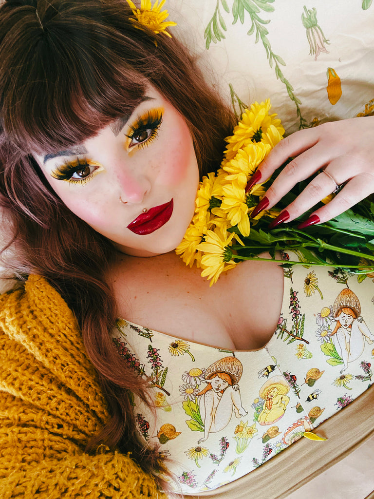 Model with yellow eyeshadow, red blush and lips and yellow flowers in their hair wearing Wilde Mode x Mushroom Babes Cottage Core Springtime Comfort Bra paired with a mustard coloured cardigan. The print is of various mythical mushroom people surrounded by a variety of plants, flowers and insects. The band on the bra is a mustard colour.